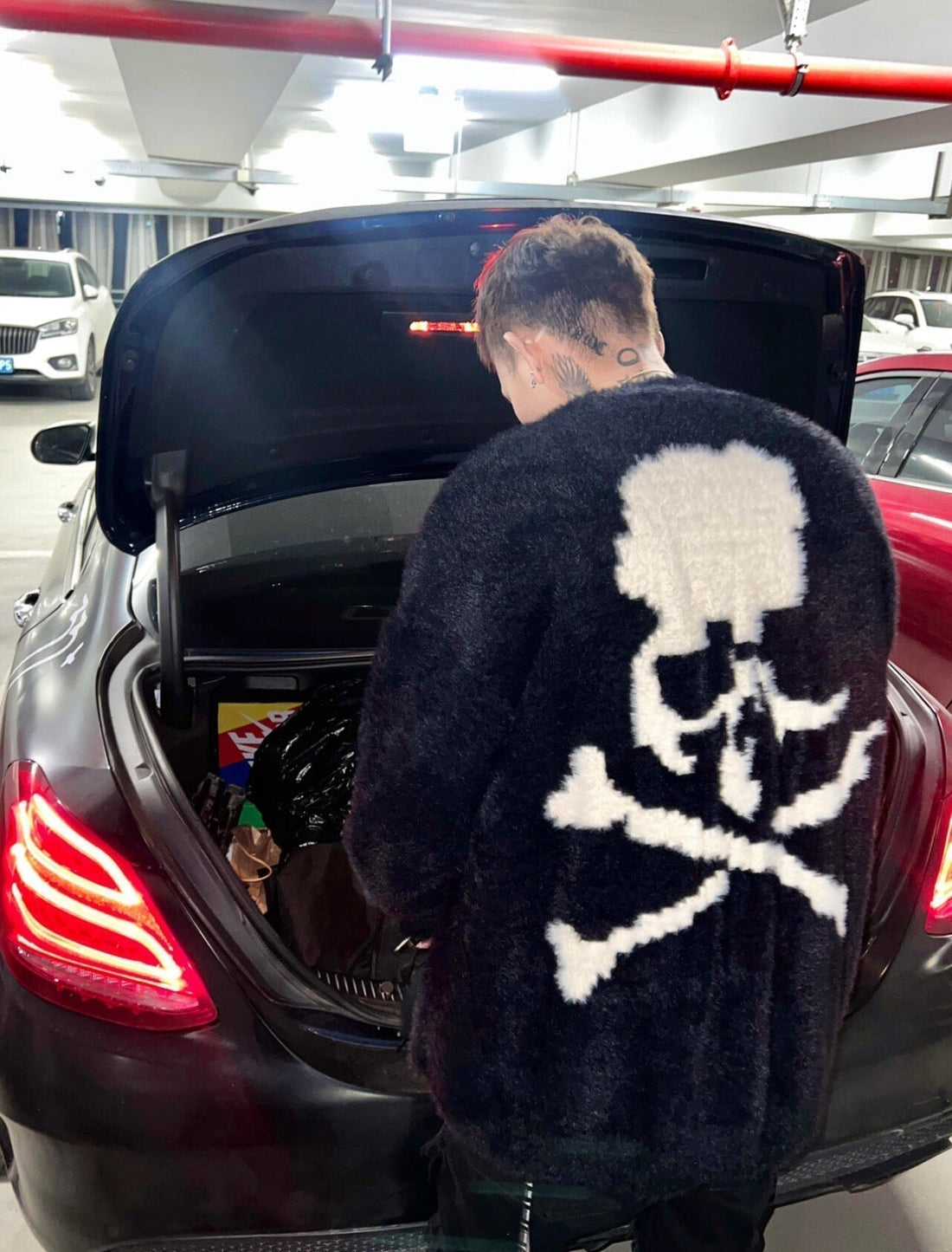 Streetwear enthusiast in fuzzy skull and crossbones jacket at car trunk, donclickin style