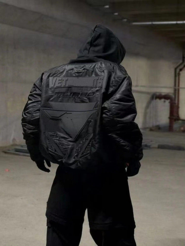 Person in black streetwear bomber jacket with bold embossed graphics in urban setting
